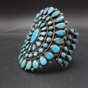 May include: A silver cuff bracelet with a large turquoise stone design. The bracelet is made of sterling silver and features a cluster of turquoise stones in a circular pattern.