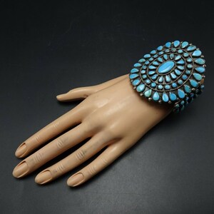 May include: A silver and turquoise bracelet with a large, circular design. The bracelet is made of sterling silver and features a cluster of turquoise stones in a sunburst pattern.