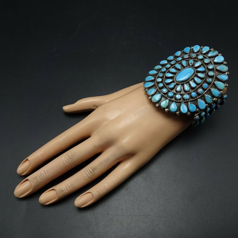 May include: A silver and turquoise bracelet with a large, circular design. The bracelet is made of sterling silver and features a cluster of turquoise stones in a sunburst pattern.