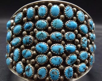 May include: A silver cuff bracelet with a wide band set with turquoise stones. The stones are oval shaped and have a dark matrix. The bracelet is made in a Native American style.