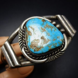 May include: A silver cuff bracelet featuring a large turquoise stone set in the centre. The stone is blue and green in colour with brown veins running through it. The bracelet has a simple design with a smooth finish.