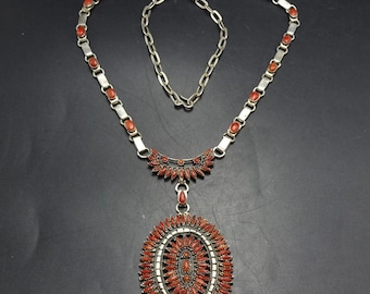 Op de afbeelding: Een zilverkleurige ketting met een grote ovale hanger en een ketting. De hanger is versierd met talrijke kleine, helderrode stenen, gerangschikt in een radiaal patroon. De ketting heeft ook rode stenen langs de ketting.
