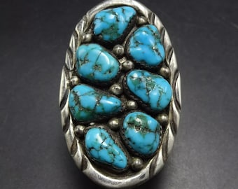 May include: A silver ring with a large oval turquoise stone set in the center. The stone is surrounded by smaller turquoise stones and silver beads.
