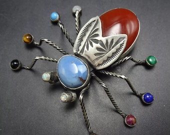 Puede incluir: Un broche de plata y piedras preciosas en forma de escarabajo con una piedra de cornalina roja, una turquesa azul y otras piedras de colores. El escarabajo tiene detalles de plata y patas de alambre retorcido.