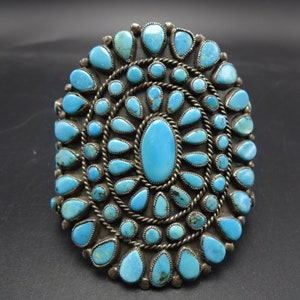 May include: A silver and turquoise Native American bracelet with a large oval turquoise stone in the center surrounded by smaller turquoise stones.