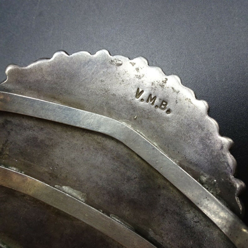 May include: Close-up of the back of a silver bracelet with a scalloped edge. The bracelet is stamped with the initials "V.M.B."