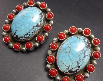 Peut inclure: Une paire de boucles d'oreilles ovales avec une pierre centrale en turquoise bleu clair à motifs matriciels sombres, entourée d'une bordure de petits cabochons ronds en corail rouge vif sertis dans une monture en métal argenté avec des perles.