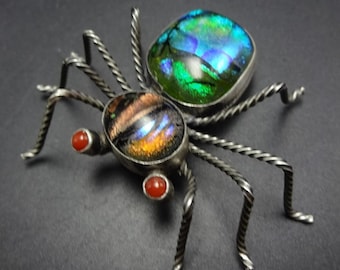 Puede incluir: Un broche de araña de plata con alas de vidrio iridiscente verde y naranja y ojos de vidrio rojo.