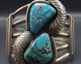 May include: A silver bracelet with two turquoise stones set in a leaf design. The bracelet is made of multiple bands of silver.