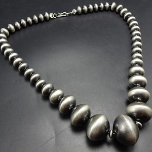 Op de afbeelding: Een zilverkleurige ketting met afgestudeerde, ronde kralen. De kralen worden groter naar het midden toe, met grotere, ovaalvormige kralen in het middelpunt. De ketting wordt getoond tegen een donkere achtergrond.