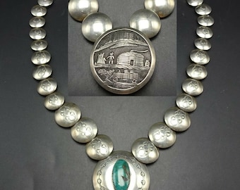 May include: A silver necklace with a large, round pendant featuring a turquoise stone. The pendant is surrounded by a decorative border. The necklace is made up of smaller, round silver beads.