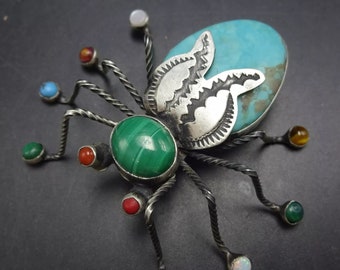 May include: A silver spider brooch with turquoise, malachite, and other gemstones. The spider has eight legs and a body with a turquoise stone.