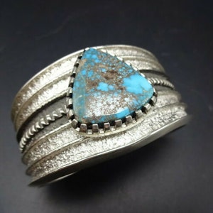 May include: A silver cuff bracelet with a turquoise stone set in a triangular shape. The stone has a blue and green color with brown and white markings. The bracelet has a textured design.