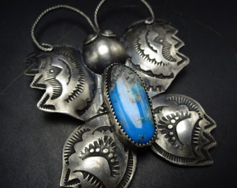 Puede incluir: Un colgante de mariposa de plata con una piedra turquesa azul en el centro. Las alas de la mariposa están detalladas con diseños intrincados.