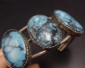 May include: A silver cuff bracelet featuring three oval-shaped turquoise stones. The stones have a blue and black marbled pattern.