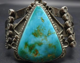 May include: A silver cuff bracelet featuring a large, triangular turquoise stone. The turquoise has green and brown veining. The bracelet has decorative silverwork around the stone and on the sides, with floral-like accents.