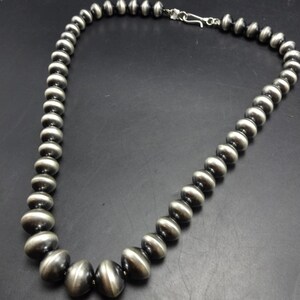 Op de afbeelding: Een zilveren kralenketting met een haaksluiting. De kralen zijn rond en in grootte afgestudeerd, met de grootste kralen aan de onderkant. De ketting wordt gepresenteerd tegen een donkere achtergrond.