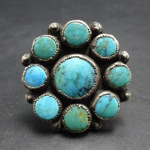 May include: A circular silver ring featuring a large central turquoise cabochon surrounded by eight smaller, round turquoise stones. The metal setting has a dark, oxidized finish with decorative silver bead accents between the stones.