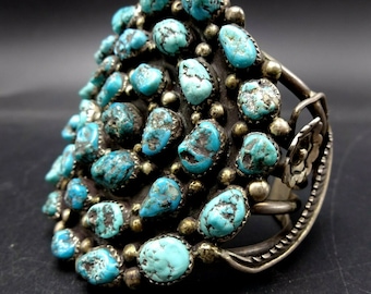 May include: A silver bracelet featuring a large cluster of turquoise stones. The stones are set in a circular pattern and are surrounded by smaller silver beads.