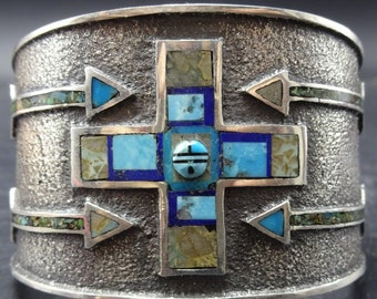 May include: A wide silver cuff bracelet with a textured surface. The bracelet features a cross design inlaid with turquoise, lapis lazuli, and other stones. Arrows are placed on the sides of the cross. The bracelet is a piece of jewelry.