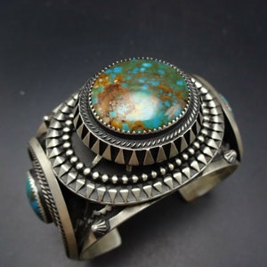 May include: A silver and turquoise cuff bracelet featuring a large, round turquoise stone at its centre. The bracelet is adorned with intricate silverwork, including beading and triangular patterns.