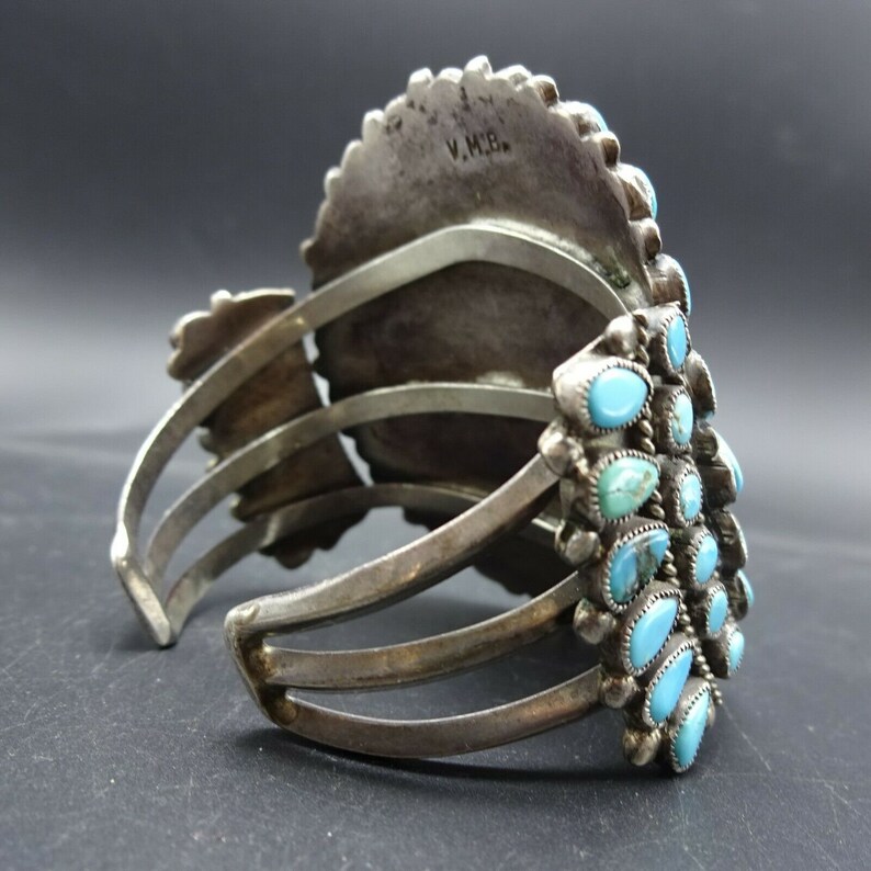 May include: A silver and turquoise bracelet with a large, round turquoise stone in the center. The bracelet has a hinged clasp and is decorated with smaller turquoise stones. The back of the bracelet is stamped with the initials "V.M.B."