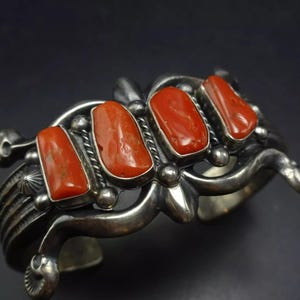 Puede incluir: Un brazalete de plata con un diseño decorativo que presenta cuatro piedras de coral rojo. El brazalete está hecho por artesanos nativos americanos.
