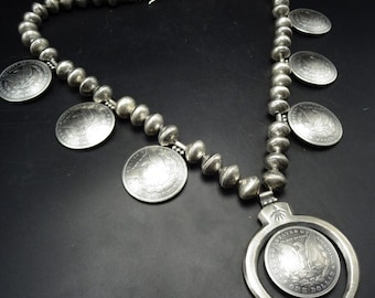 May include: A silver necklace with a large silver pendant shaped like a horseshoe. The pendant has a silver coin with the words "United States of America" and "One Dollar" inscribed on it. The necklace is made of silver beads and has several silver coins attached to it.