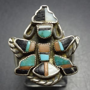 May include: A silver-toned ring with a unique, geometric design. The ring features inlaid stones in black, white, turquoise, and brown. The ring's design is symmetrical, with a central cluster of stones and surrounding decorative elements.