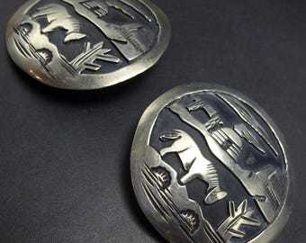 Puede incluir: Dos hebillas de cinturón redondas plateadas con un diseño en relieve detallado. Cada hebilla presenta una imagen central de un animal parecido a un caballo, rodeado de motivos estilizados de montañas y plantas. El fondo es oscuro, realzando los detalles plateados.