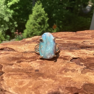 May include: A silver ring featuring a large, oval-shaped gemstone with a mix of blue, white, and pale pink hues. The gemstone is set with four prongs, and the ring is displayed on a piece of textured, brown wood, with a blurred green background.
