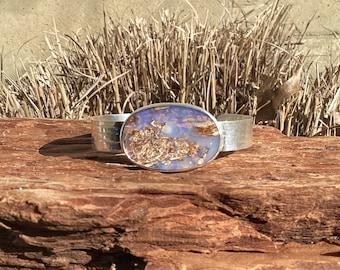 May include: Silver cuff bracelet with an oval, multi-colored stone. The stone features shades of blue, white, and brown. The silver band has a textured design. The bracelet is displayed on a piece of wood.