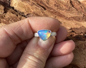 May include: A silver ring featuring a triangular opal gemstone with iridescent blue, yellow, and green hues. The ring band has a textured, patterned design. The opal is the focal point, showcasing a play of color.