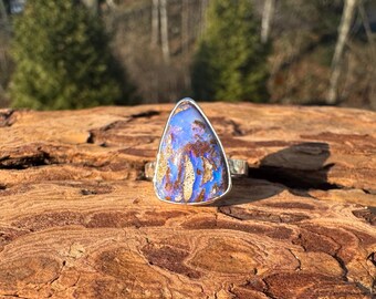 May include: A silver ring with a large, triangular opal stone. The opal has a blue and gold color pattern. The ring is set on a textured, brown surface, with a blurred background of trees and sunlight.