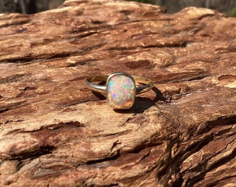 May include: A gold ring with a rectangular opal gemstone. The opal displays a vibrant play of color, including hues of orange, green, and blue. The ring is set on a textured, brown wooden surface.