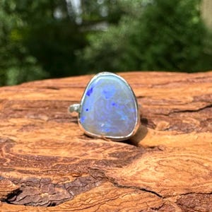 May include: A silver ring featuring a large, oval-shaped opal stone. The opal displays a mix of blue and white colors with a shimmering effect. The ring is set on a piece of textured, brown wood.