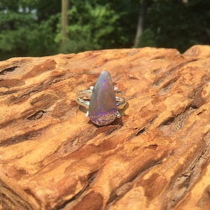 Peut inclure: Bague en argent ornée d'une pierre en forme de goutte d'eau. La pierre présente une base gris foncé avec des éclats violets et dorés. La bague est posée sur une surface en bois brun texturé, avec un arrière-plan vert flou.
