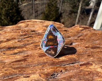 May include: A silver ring featuring a large, teardrop-shaped opal. The opal displays a vibrant array of colors, including blue, pink, and gold, set against a dark brown matrix. The ring is displayed on a textured, brown wooden surface.