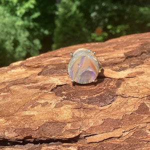 May include: A pear-shaped, iridescent gemstone ring with a silver band. The gemstone displays a mix of colors, including orange, purple, and yellow. The ring is set against a natural wood background, with green foliage visible in the distance.