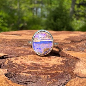 Op de afbeelding: Zilveren ring met een ovale, veelkleurige steen. De steen heeft paarse, blauwe en bruine tinten. De ring is gezet in een zilveren bezel en wordt gepresenteerd op een getextureerd, bruin houten oppervlak.