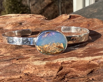 May include: A silver cuff bracelet with a hammered texture. The centerpiece is an oval-shaped stone with a landscape design in shades of blue, brown, and white. The bracelet is displayed on a piece of wood.