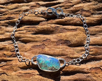 May include: A silver bracelet featuring a vibrant, multi-colored opal stone. The opal is set in a silver bezel and connected to a delicate chain. The bracelet is displayed on a textured, brown wooden surface.
