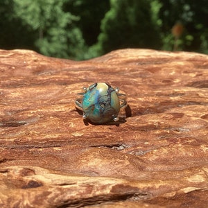 May include: A silver ring featuring a large, oval-shaped stone with a unique pattern of blue, green, and brown hues. The ring's setting has a delicate, open design, with small silver accents around the stone. The ring is displayed on a textured, wooden surface.