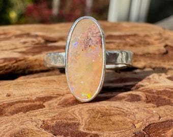 May include: A silver ring with an oval-shaped opal stone. The opal has a mix of colors, including yellow, orange, and hints of blue. The ring's band has a textured design. The ring is set on a wooden surface.