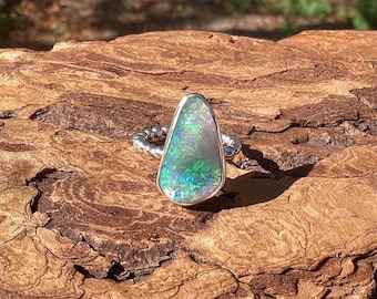 May include: A silver ring featuring a large, pear-shaped opal with iridescent green and blue hues. The opal is set in a polished silver bezel, and the band has a textured, beaded design. The ring is displayed on a textured, brown wooden surface.