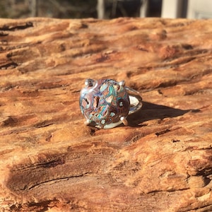 May include: A silver ring with a unique, colorful stone. The stone features a mosaic of brown, teal, and blue hues. The ring is set against a textured, brown wooden surface, creating a striking contrast.