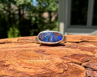 May include: A silver ring featuring a large, oval-shaped opal. The opal displays a vibrant play of color, with flashes of blue, green, and purple. The ring's band is a simple, polished silver, highlighting the gemstone's beauty.