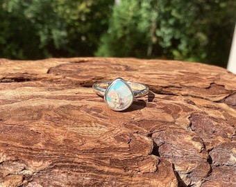 May include: A silver ring with a pear-shaped opal gemstone. The opal displays a mix of colors, including blue, green, and brown. The ring is set on a textured wooden surface, with a blurred green background.