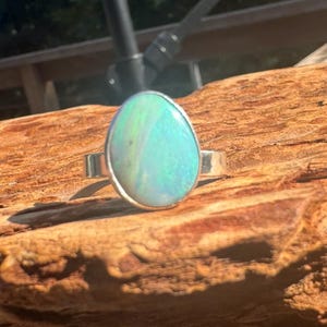 May include: A silver ring featuring a large, oval opal gemstone. The opal displays a vibrant play of color, with hues of blue, green, and hints of yellow. The ring is set on a natural wood surface.
