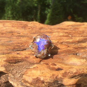 May include: A silver ring featuring a large, oval-shaped opal gemstone. The opal displays a mix of brown, tan, and blue hues, with a prominent flash of vibrant blue. The ring is set on a natural wooden surface.